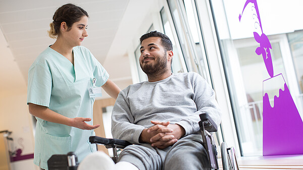 Ein Mann im Rollstuhl und eingegipstem Bein schaut eine Krankenpflegerin an, die links neben ihm steht und ihn anschaut.