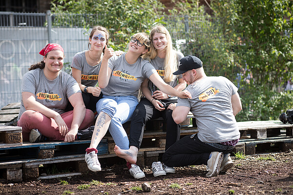 Eine Gruppe junger Menschen sitzt auf Holzpaletten zusammen und lächelt in die Kamera.