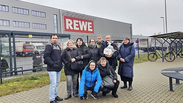 Eine Gruppe von Menschen steht vor einem Lagergebäude von REWE. In der hinteren Reihe stehen sieben Menschen, davor sind zwei Menschen in der Hocke. 