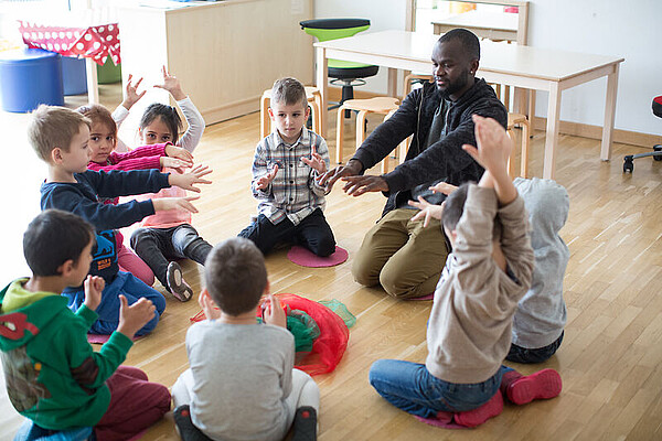 Acht Kinder sitzen zusammen mit einem Erzieher im Kreis auf dem Boden.