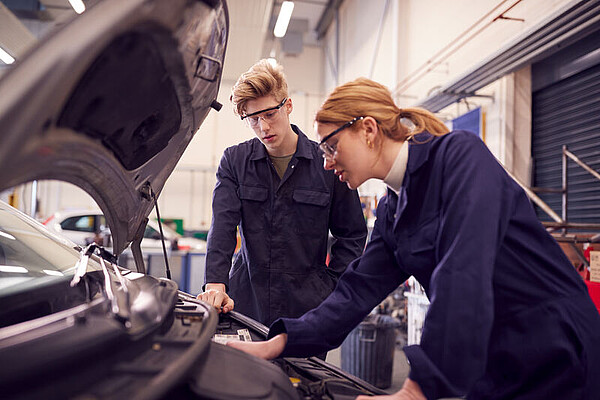 Zwei junge Personen in einer KFZ-Werkstatt schauen in die Motorhaube eines Autos.