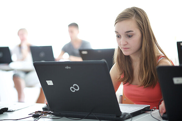 Junge Frau sitzt an einem Tisch vor einem Laptop. Im Hintergrund sind zwei weitere Personen zu sehen.