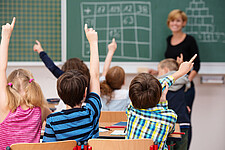 Schulkinder in einem Klassenraum, vorne an der Tafel steht eine Lehrerin. Mehrere Kinder melden sich.