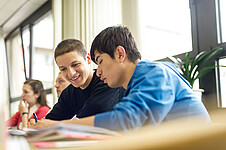 Zwei Schüler in einem Klassenzimmer, der eine lächelt, der andere hält einen Kugelschreiber. Im Hintergrund sind weitere Personen zu sehen.