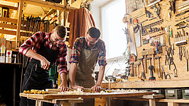 Zwei Männer arbeiten in einer Werkstatt mit einer Bohrmaschine und Holz.
